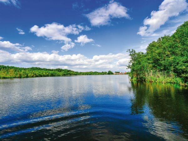 Seenlandschaft im Land Brandenburg mit bewaldeten Ufern und einem Dorf am See. Weiße Wolken ziehen über den blauen Himmel.