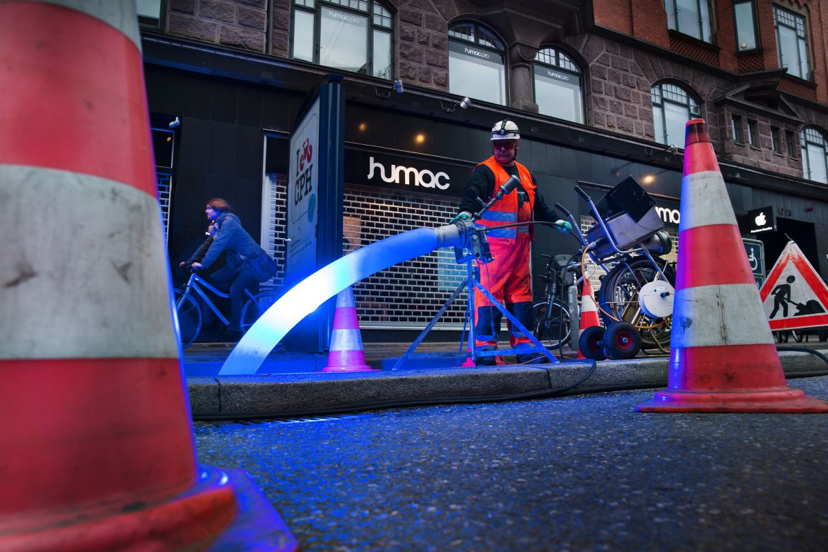 Ein Kanalarbeiter mit Helm und Schutzkleidung bedient einen LED-Strahler für die Lichthärtung eines Schlauchliners auf einer Baustelle. Die Baustelle, die sich an einer Geschäftsstraße befindet, ist mit Pylonen und Baustellenschild abgesichert. Eine Radfa