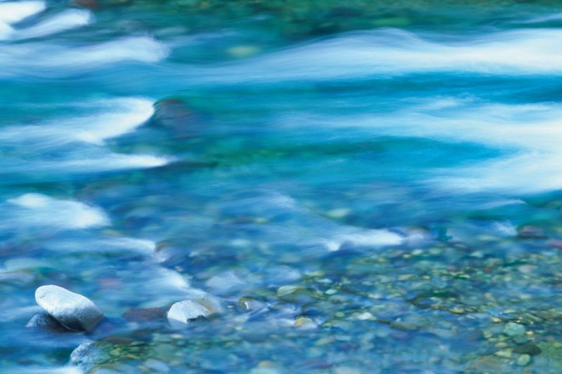 "Blauweißes Wasser" fließt in einem Bach in flachen Wellen über das steinige Bachbett. Ein grüner Uferstreifen ist schemenhaft zu sehen.