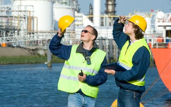 2 Hafenarbeiter mit gelben Schutzhelmen und Warnwesten schauen hoch in die gleiche Richtung. Sie stehen in der Nähe eines ankernden Schiffes und am gegenüberliegenden Ufer befindet sich eine Ölraffenerie.