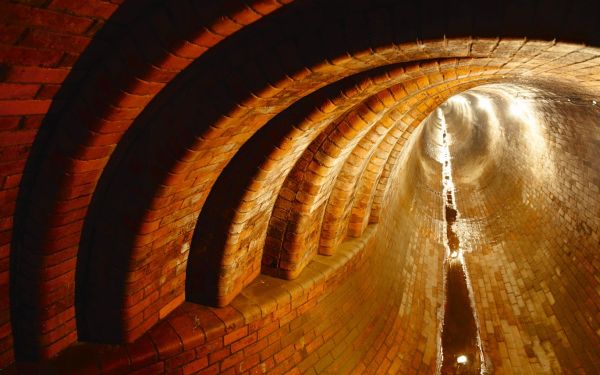 Vollständig mit Backsteinen ausgekleidetes Kurven-Teilstück einer unterirdichen Kanalisation mit Beleuchtung, führt wenig Abwasser.