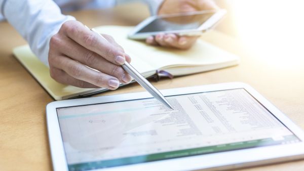 Eine Person prüft eine Abrechnung auf einem Tablet-Computer. Es ist eine Nahaufnahme der Hände der Person zu sehen, die einen Kuli und ein Handy halten. Ein Notizbuch ist aufgeschlagen. Davor liegt das Tablet.