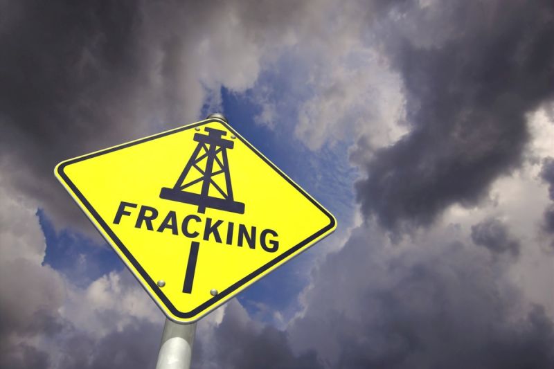 gelbes Schild mit der Aufschrift FRACKING und einer mit Förderturm-Zeichnung, Hintergrund ein Himmel mit Gewitterwolken