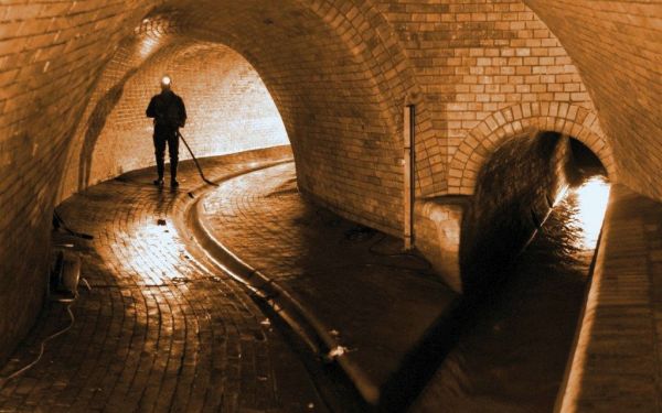 Ein Kanalarbeiter in Gummistiefeln und mit Stirnlampe reinigt mit einer Schaufel eine Rinne in der Kanalisation in der Nähe einer Einmündung eines kleineren Kanals. Eine Stromkabelrolle steht an der linken Wand und ist teilweise abgerollt.