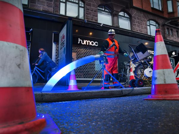 Ein Kanalarbeiter mit Helm und Schutzkleidung bedient einen LED-Strahler für die Lichthärtung eines Schlauchliners auf einer Baustelle. Die Baustelle, die sich an einer Geschäftsstraße befindet, ist mit Pylonen und Baustellenschild abgesichert. Eine Radfa