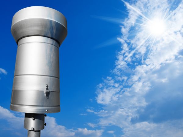 Wetterstation mit einem Regenmesser aus Metall vor einem blauen Himmel mit weissen Wolken von der sonne durchschienen.