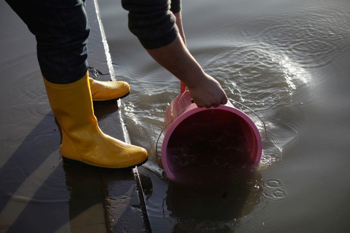 Mann mit gelben Gummi-stiefeln schöpft Schmutzwasser durch Überflutung bei Hochwasser, mit einem rosafarbenen Eimer.