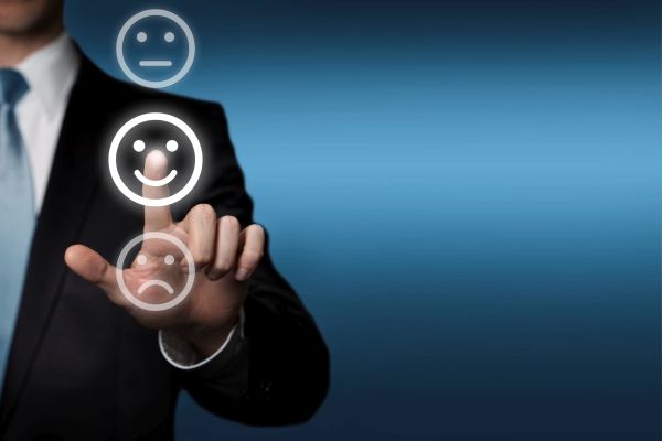 Geschäftsmann steht an einer transparenten Touchscreen-Fläche. Er berührt mit seinem Zeigefinger das mittlere, lächelnde, der 3 Smile-Symbole.