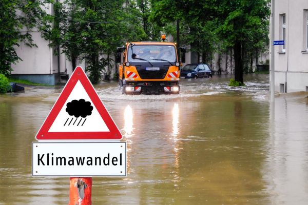 Im Hochwasser steht ein dreieckiges Warnschild mit einer schwarzen Wolke, aus der es schwarz regnet und einem Schild darunter mit dem Schriftzug "Klimawandel". Im Hintergrund fährt ein Nutzfahrzeug durchs Hochwasser und ein kleiner PKW steht am überflutet