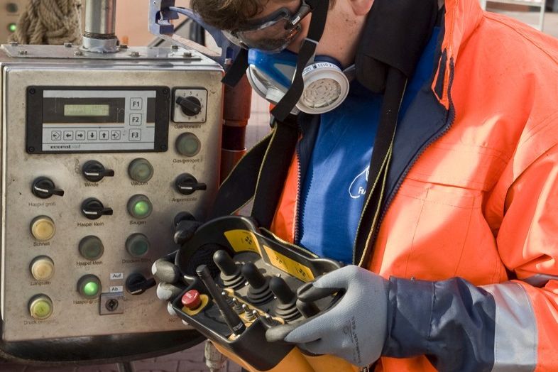 Auf einem gepflasterten Aussenbereich bedient ein Arbeiter in Schutzkleidung ein Fernsteuerungsgerät, daß an einem Band um den Hals hängt.