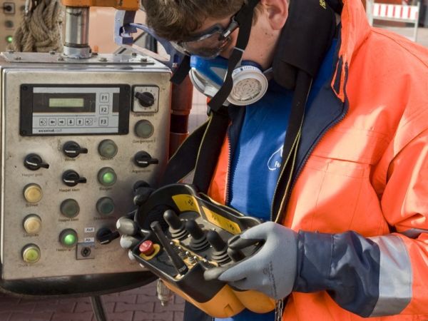 Auf einem gepflasterten Aussenbereich bedient ein Arbeiter in Schutzkleidung ein Fernsteuerungsgerät, daß an einem Band um den Hals hängt.