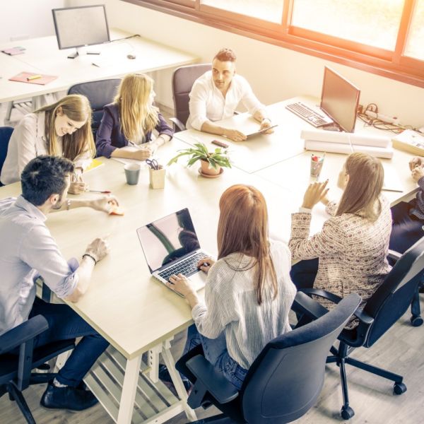 unges Mitarbeiterteam sitzt zusammen an einem großen Tisch und diskutiert. 3 Frauen schreiben mit Stift oder am Laptop. Ein Mann hält ein Tablet in den Händen.