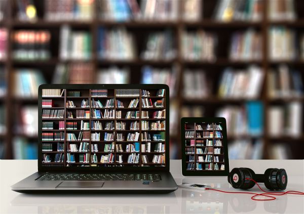 Auf einer glänzenden Tischoberfläche befinden sich ein aufgeklapptes Laptop, ein Tablet und Kopfhörer. Die Bildschirme zeigen ein Bücherregal einer Bibliothek, das gleichzeitig den unscharfen Hintergrund bildet.