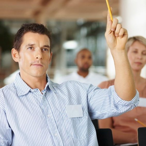 Ein Journalist meldet sich mit einem gelben erhobenen Stift in der linken Hand zu Wort. Noch andere Journalist*innen sitzen hinter ihm und machen sich Notizen.