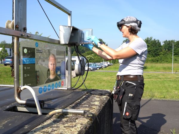 Mitarbeiterin mit Hololensbrille steht vor einem Kasten. Links davon ist das Bild von einem anderen Mitarbeiter zu sehen, was von der Hololens übertragen wird