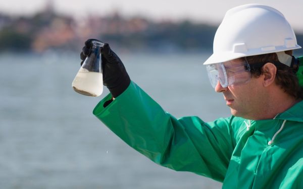 Umweltinspektor, mit Schutzbrille und weissem Schutzhelm, der eine Wasserprobe in einem Erlenmeyerkolben/Glas am Entnahmeort visuell überprüft