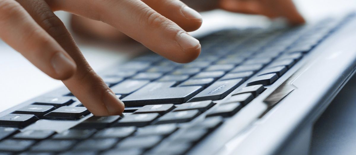 Eine Person bedient zweihändig eine Computertastatur, wobei die linke Hand und der größte Teil der Tastatur undeutlich zu sehen ist und die rechte Hand im Vordergrund deutlich.