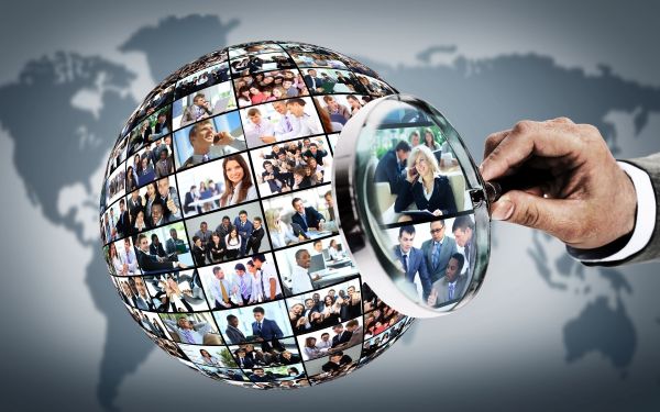 Eine Hand hält eine Lupe auf Fotos mit Menschen gerichtet. Auf den Fotos befinden sich meist Teams von Menschen, aber auch Einzelpersonen, die im Geschäftsleben stehen. Die Fotos sind dicht nebeneinander auf einer Kugel angeordnet. Im Hintergrund ist die