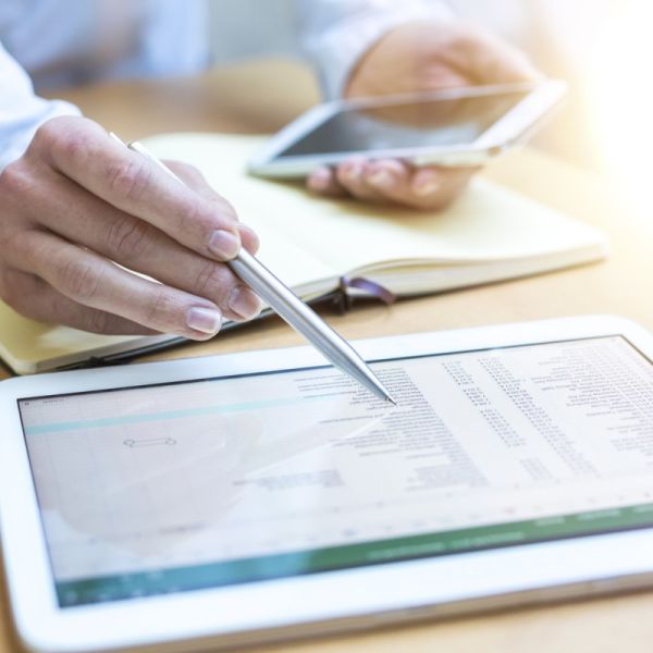 Eine Person prüft eine Abrechnung auf einem Tablet-Computer. Es ist eine Nahaufnahme der Hände der Person zu sehen, die einen Kuli und ein Handy halten. Ein Notizbuch ist aufgeschlagen. Davor liegt das Tablet.