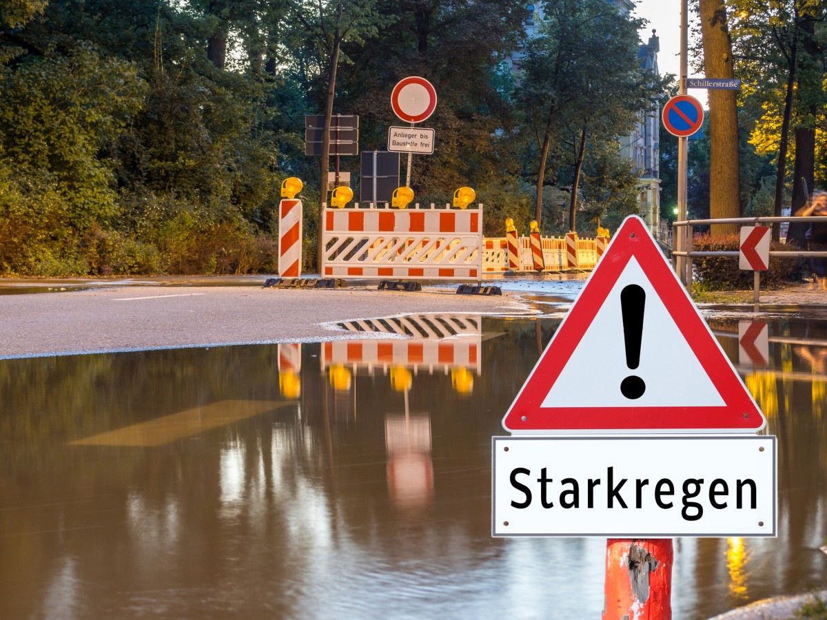 Überflutung u.a. einer Schillerstraße durch Starkregen. Achtung Starkregen Warnschild! Baustellenschild mit "Anlieger bis Baustelle frei" Einschränkungshinweisschild, umgeben von Absperrzäunen und -baken z.T. mit Warnbeleuchtung in einer Stadt.