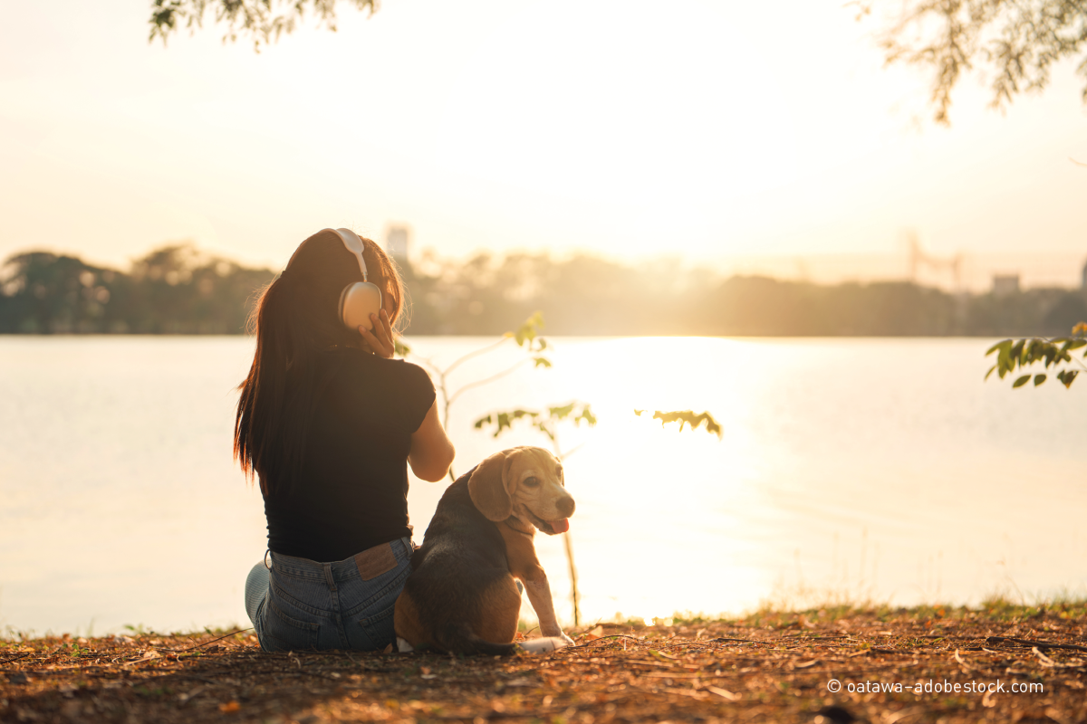 Mädchen mit Kopfhörern sitz mit ihrem Hund am Flussufer in der untergehenden Sonne