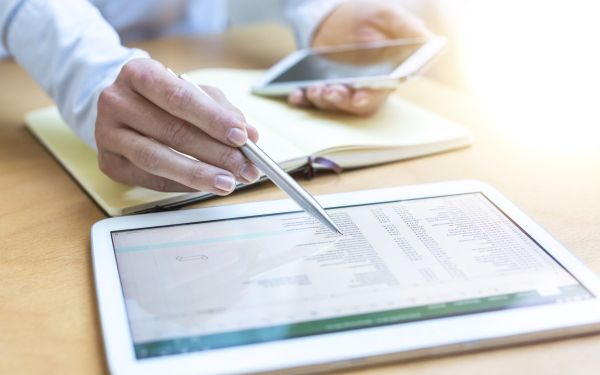 Eine Person prüft eine Abrechnung auf einem Tablet-Computer. Es ist eine Nahaufnahme der Hände der Person zu sehen, die einen Kuli und ein Handy halten. Ein Notizbuch ist aufgeschlagen. Davor liegt das Tablet.