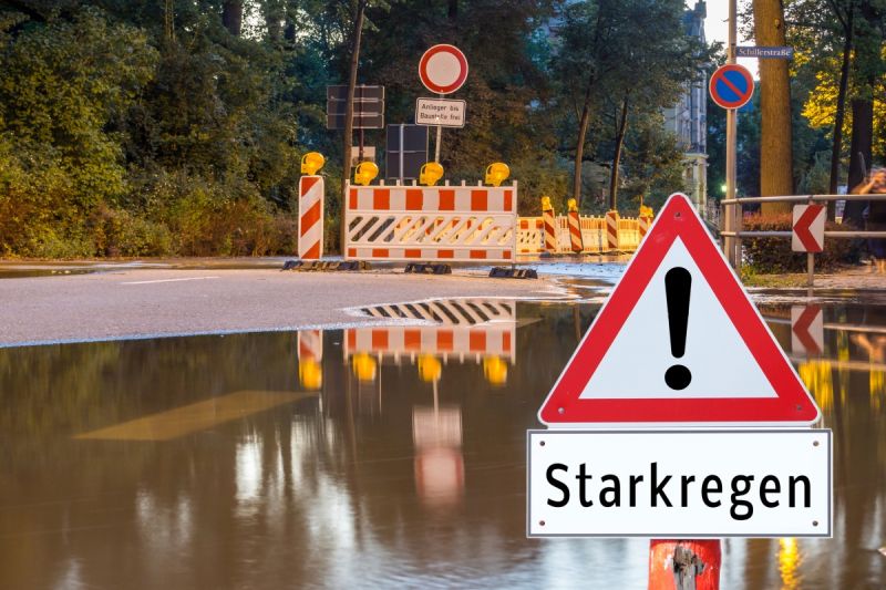 Überflutung u.a. einer Schillerstraße durch Starkregen. Achtung Starkregen Warnschild! Baustellenschild mit "Anlieger bis Baustelle frei" Einschränkungshinweisschild, umgeben von Absperrzäunen und -baken z.T. mit Warnbeleuchtung in einer Stadt.