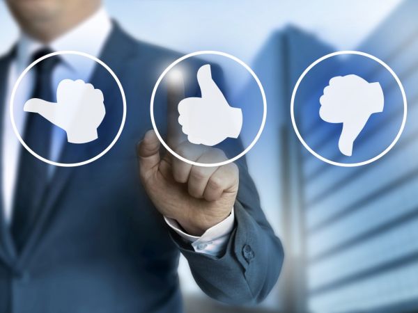 Hinter einer Glasscheibe mit 3 Emojis Buttons mit Daumen hoch, runter und nach links steht ein Geschäftsmann, der mit dem linken Zeigefinger auf den mittleren Button mit Daumen nach oben zeigt.