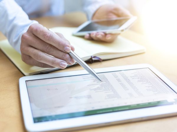 Eine Person prüft eine Abrechnung auf einem Tablet-Computer. Es ist eine Nahaufnahme der Hände der Person zu sehen, die einen Kuli und ein Handy halten. Ein Notizbuch ist aufgeschlagen. Davor liegt das Tablet.