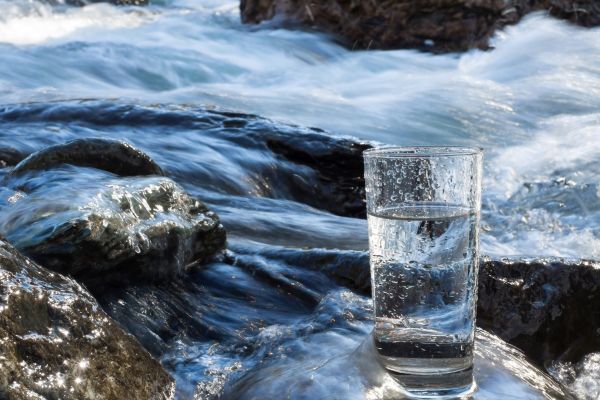Klares Wasser in einem Glas, das auf einem mit Wasser überspülten Stein steht. Er befindet sich in einem Bachlauf mit vielen Steinen.