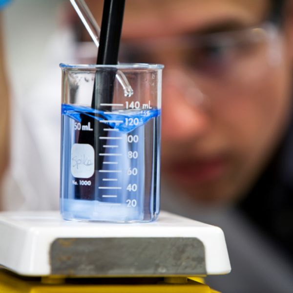 Ein Laborant mit Schutzbrille und Schutzhandschuhen tropft mit einer Pipette eine klare Füssigkeit in ein Messbecherglas mit anderer klarer Flüssigkeit, leicht blau gefärbt. Er rührt sie dabei gleichzeitig mit einem schwarzen Rührstab um. Das Becherglas s